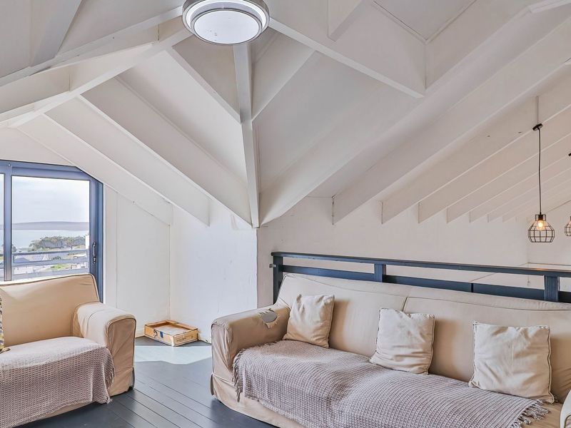 A bright living area featuring a high vaulted ceiling with exposed rafters, modern pendant lighting, and a sliding glass door offering scenic coastal views.