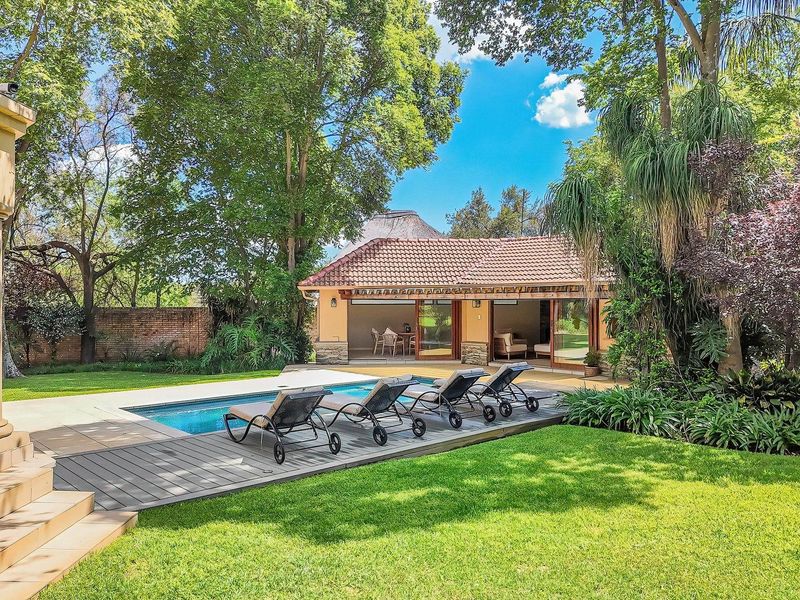 A well-maintained outdoor pool area featuring a swimming pool, wooden decking, and a landscaped garden with a secondary building in the background.