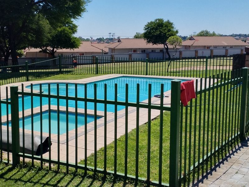 Communal swimming pool area featuring a paved surround, secure metal fencing, and well-maintained lawn within a residential townhouse complex.