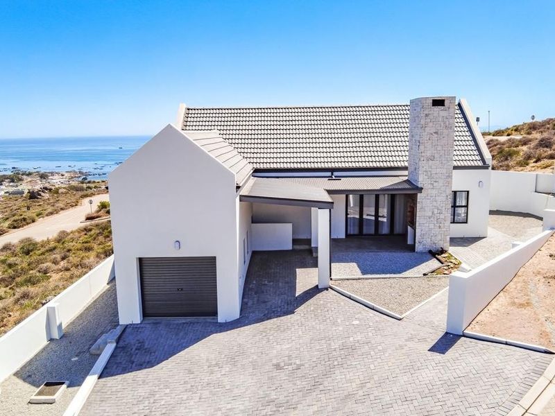 Aerial view of a modern coastal home featuring a paved driveway, single garage, stone-clad chimney, and expansive ocean views.