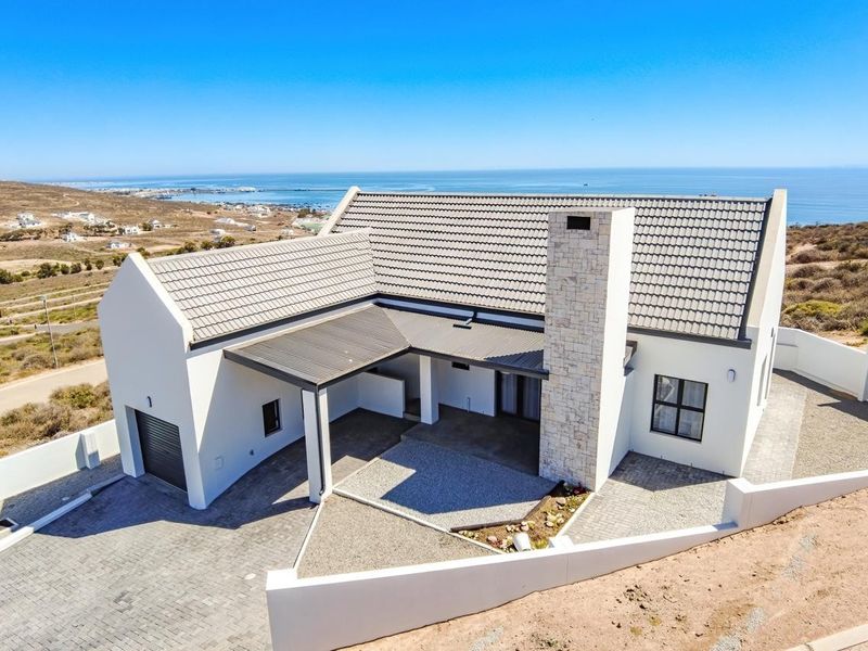 Aerial view of a modern coastal home featuring a stone-clad chimney, tiled roof, paved driveway, and a covered patio with scenic ocean views.