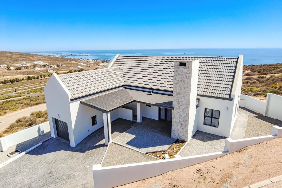 Aerial view of a modern coastal home featuring a stone-clad chimney, tiled roof, paved driveway, and a covered patio with scenic ocean views.