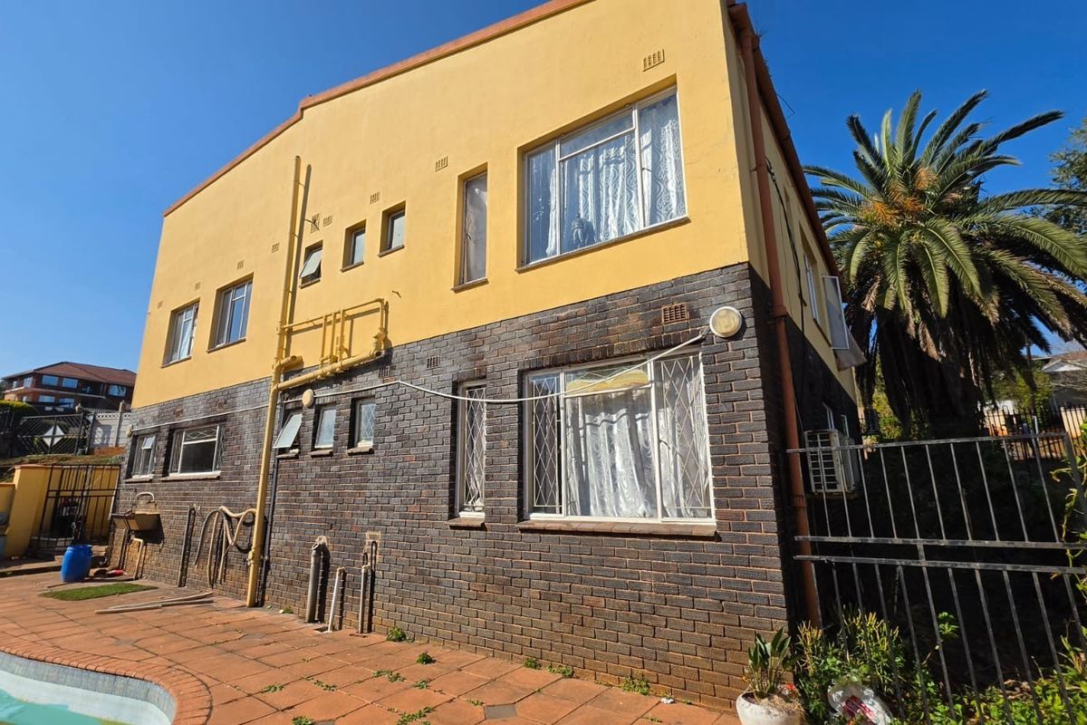 Rear elevation of a two-story house featuring a combination of brick and plastered walls, metal window frames with security bars, and an adjacent paved patio area.
