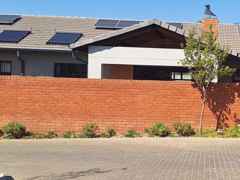 Front view of a townhouse featuring a brick boundary wall, paved driveway, and roof-mounted solar panels and solar water heating system.