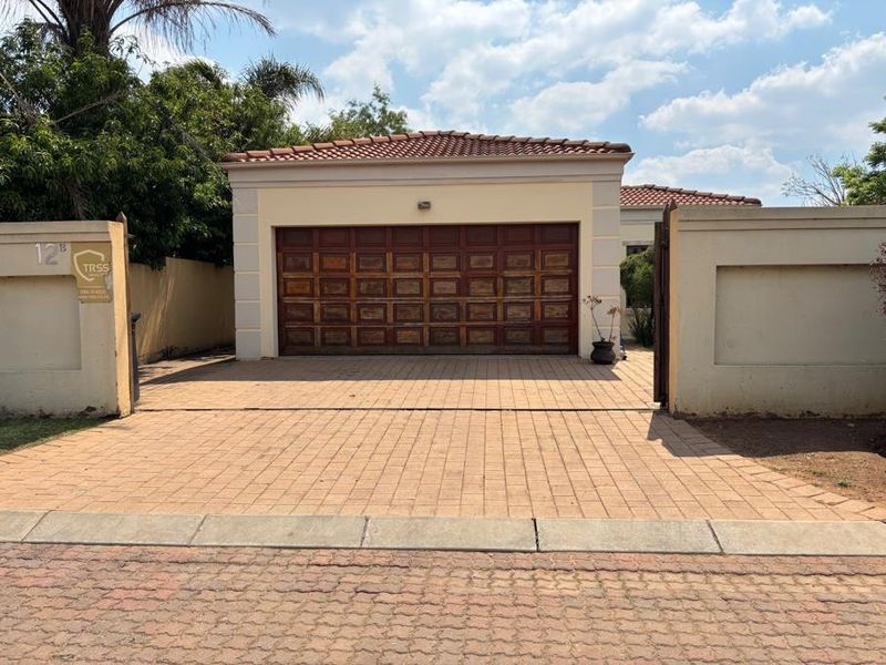 Front elevation of the property featuring a paved driveway, perimeter wall with gated entrance, and a wooden garage door under a tiled roof.