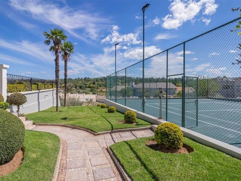 A well-maintained private tennis court featuring chain link fencing, floodlights, and an adjacent landscaped garden with a paved walkway.