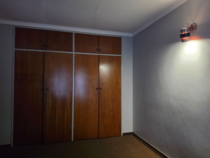 A vacant bedroom featuring built-in wooden cupboards, neutral grey walls, and carpeted flooring, illuminated by a wall-mounted light fixture.