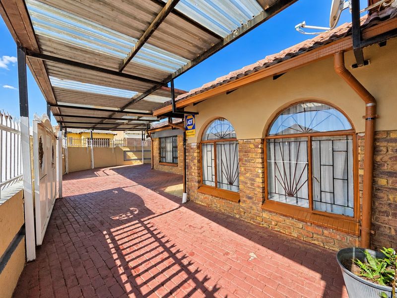 A covered outdoor patio area featuring brick paving, arched windows with security bars, and a corrugated roof structure.