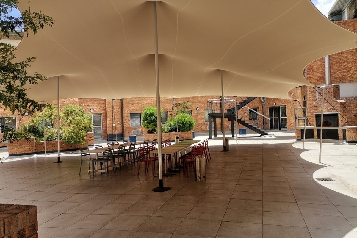 A spacious outdoor courtyard featuring a large tensile fabric canopy, tiled flooring, and integrated brick planters, suitable for commercial or social gatherings.