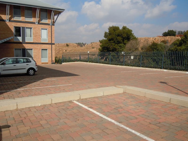A paved commercial parking area adjacent to a multi-story brick building, featuring secure metal fencing and marked parking bays.