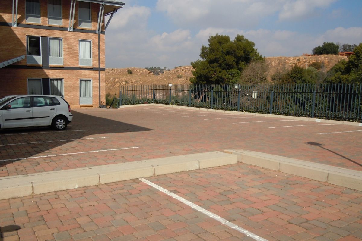 A paved commercial parking area adjacent to a multi-story brick building, featuring secure metal fencing and marked parking bays.