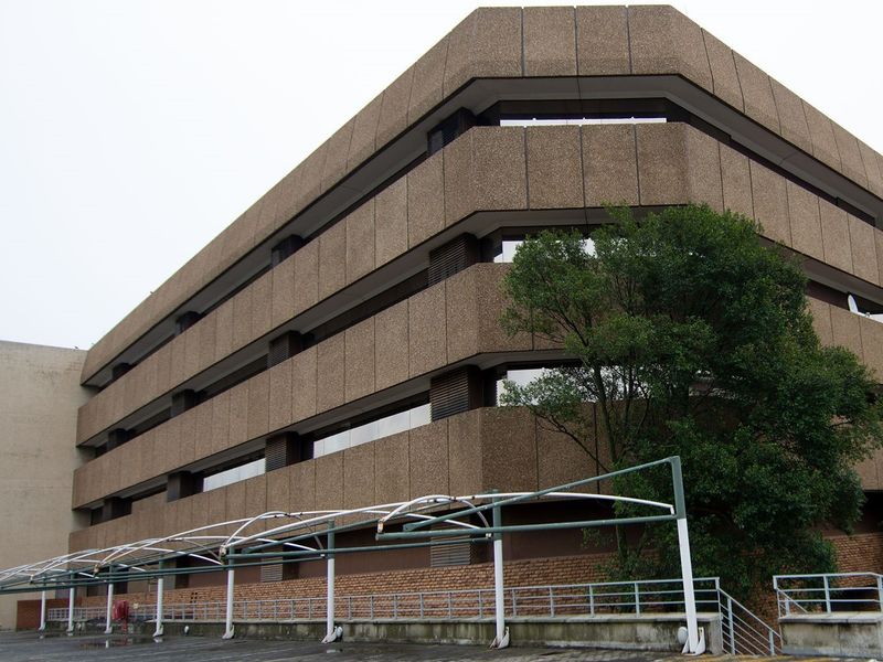 A multi-story commercial building featuring a textured concrete facade, tinted windows, and an external metal awning structure over a paved parking area.