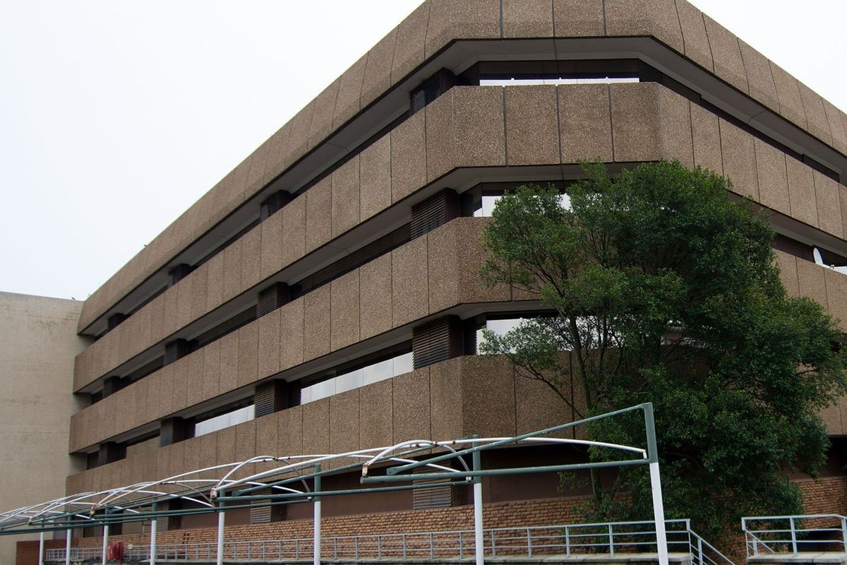 A multi-story commercial building featuring a textured concrete facade, tinted windows, and an external metal awning structure over a paved parking area.