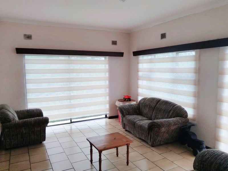 A bright living room featuring neutral tiled flooring, large windows with modern zebra blinds, and decorative cornicing.