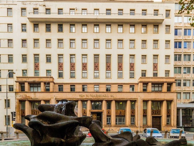 A grand commercial building facade featuring classical architectural elements, a multi-story window grid, and a prominent stone fountain in the foreground.