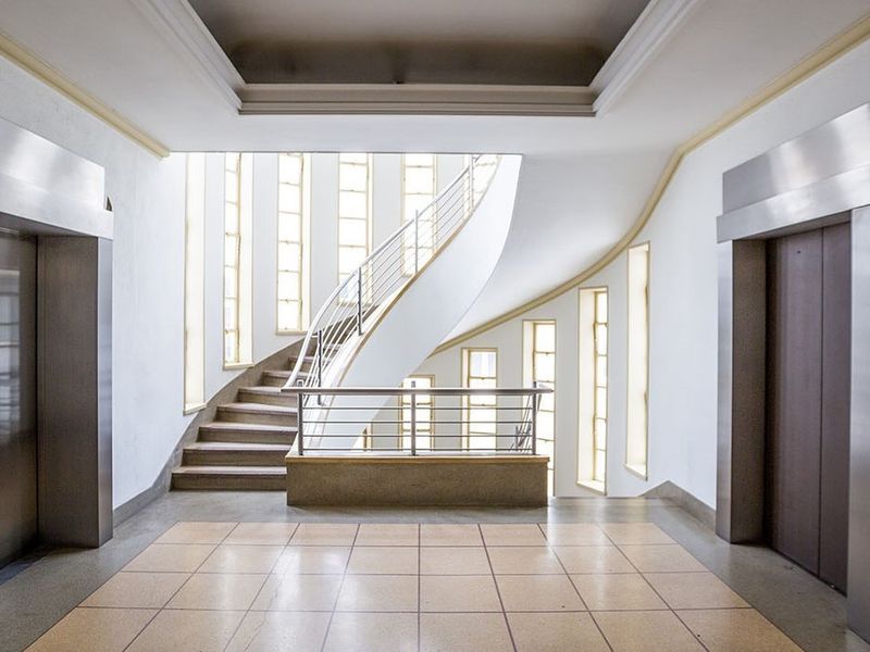 A bright, modern commercial hallway featuring a striking curved staircase, stainless steel elevator access, and large vertical windows providing ample natural light.