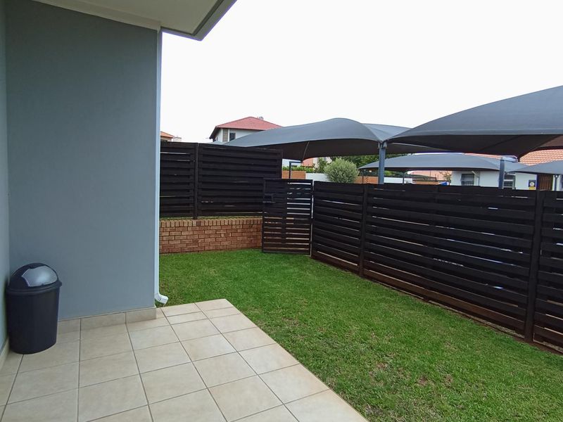 A neat outdoor patio area featuring tiled flooring, timber privacy fencing, and overhead shade sails, overlooking a small private garden space.