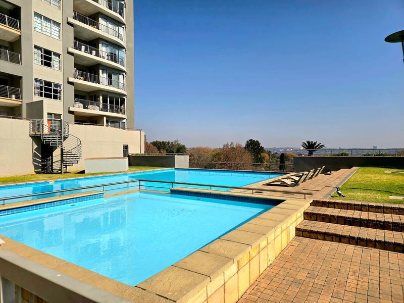 A well-maintained outdoor swimming pool area featuring a paved deck, metal safety fencing, and a spiral staircase leading to the apartment building.