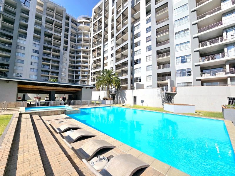 A well-maintained communal swimming pool area featuring a paved deck, integrated seating, and views of the surrounding apartment complex architecture.