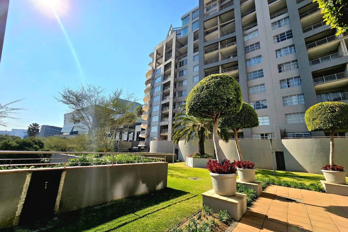 A well-maintained landscaped garden area with paved terrace and retaining walls, set against the backdrop of a multi-story apartment building.