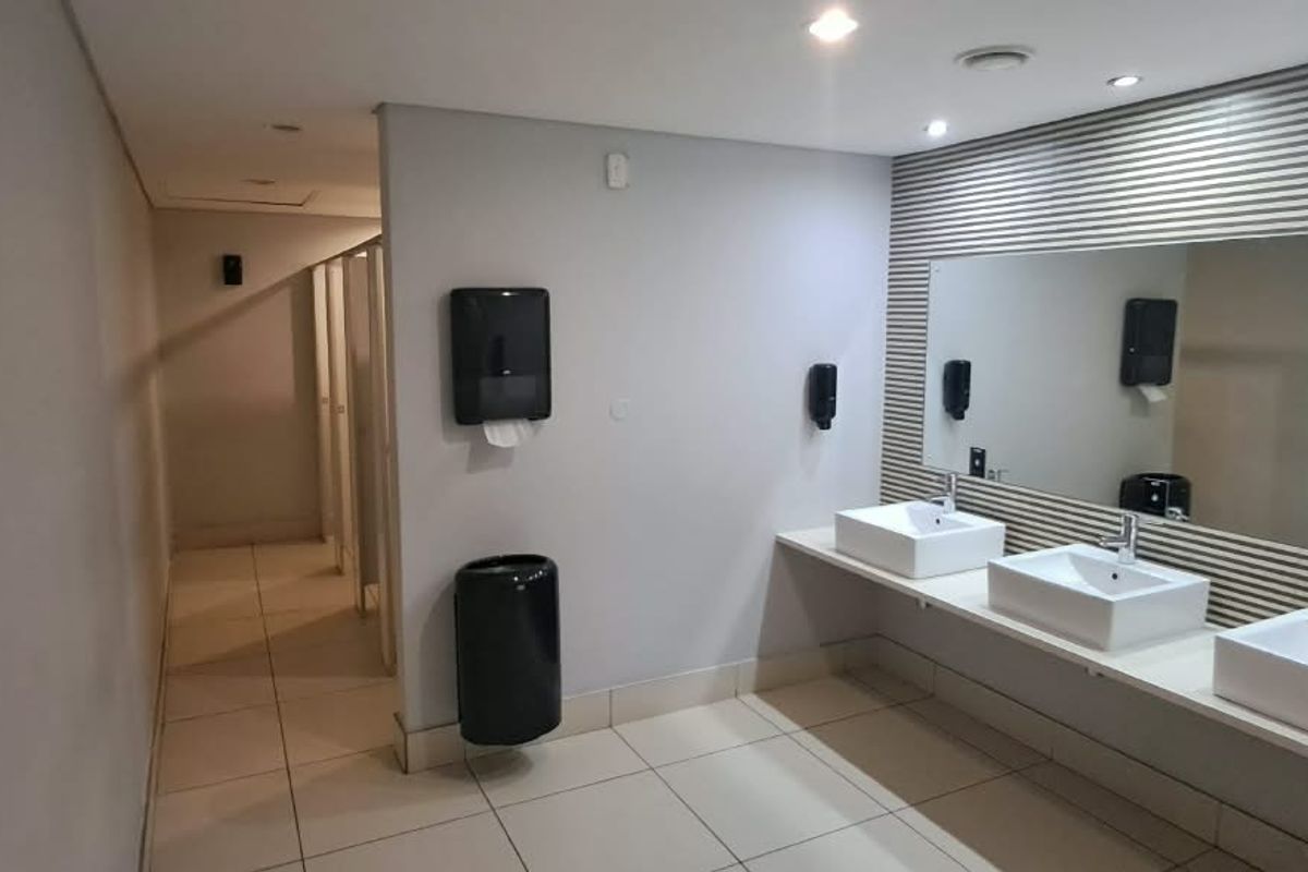 Modern office bathroom featuring a triple vanity with ceramic basins, large wall mirror, and private toilet cubicles with clean tiled flooring.