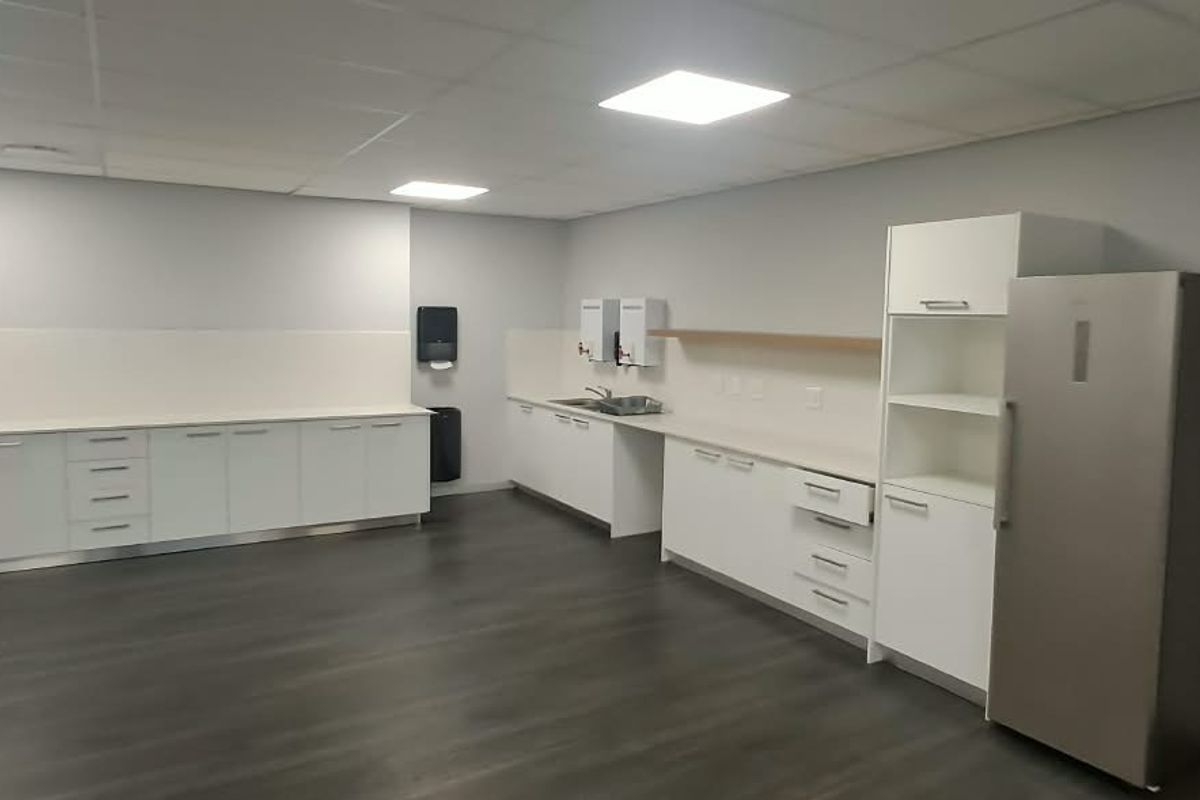 A clean, modern office kitchenette featuring white built-in cabinetry, laminate countertops, a stainless steel sink, and durable dark vinyl flooring.