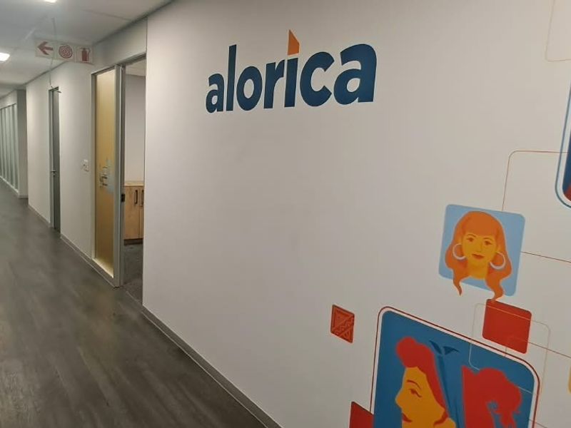A clean, modern office hallway featuring laminate flooring, recessed ceiling lights, and glass-partitioned office entrances with branded wall graphics.