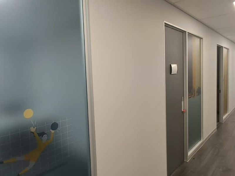 A clean, modern office hallway featuring laminate flooring, recessed ceiling lights, and frosted glass partitions leading to individual office spaces.