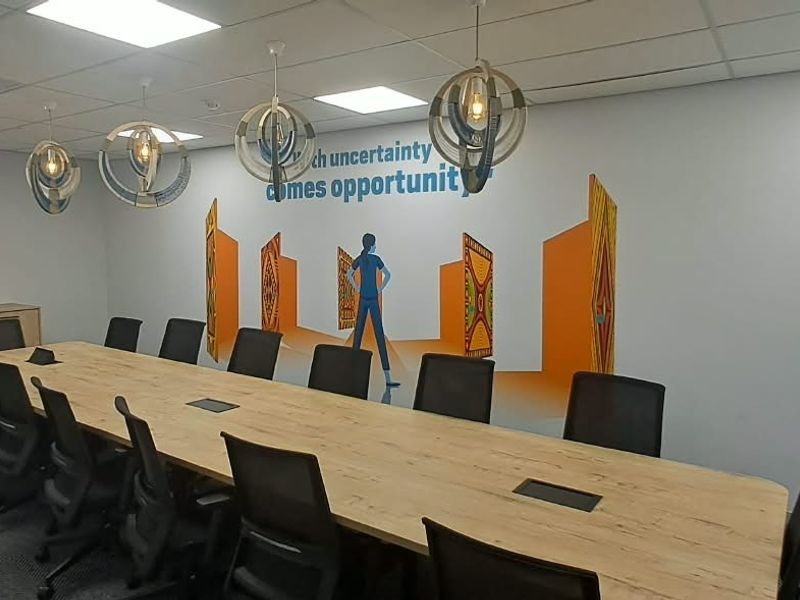 A professional office boardroom featuring a large central table, suspended decorative lighting, carpeted flooring, and a feature wall with graphic branding.