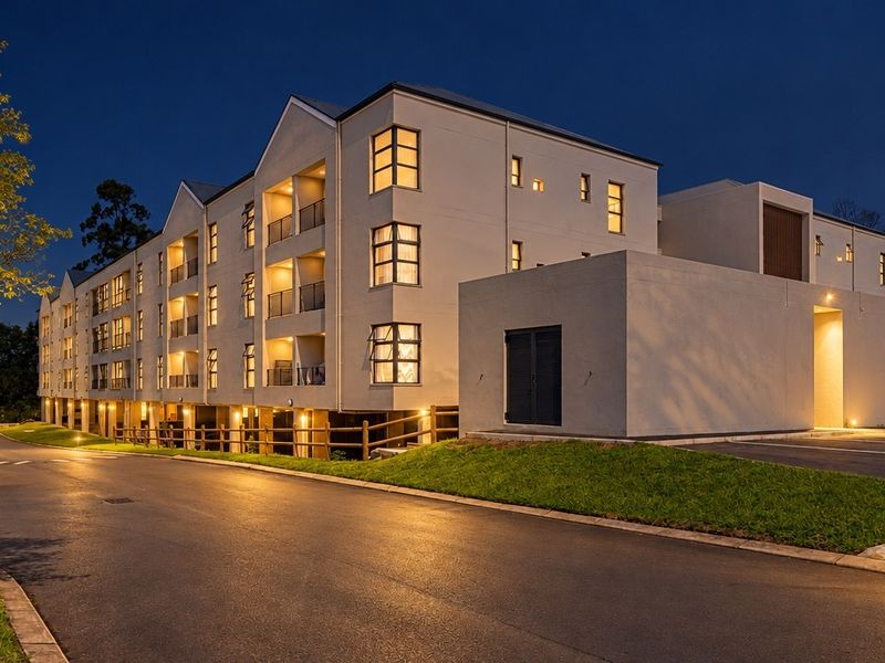 A well-maintained multi-story apartment complex at dusk, featuring modern architectural design, private balconies, and paved driveway access with integrated exterior lighting.