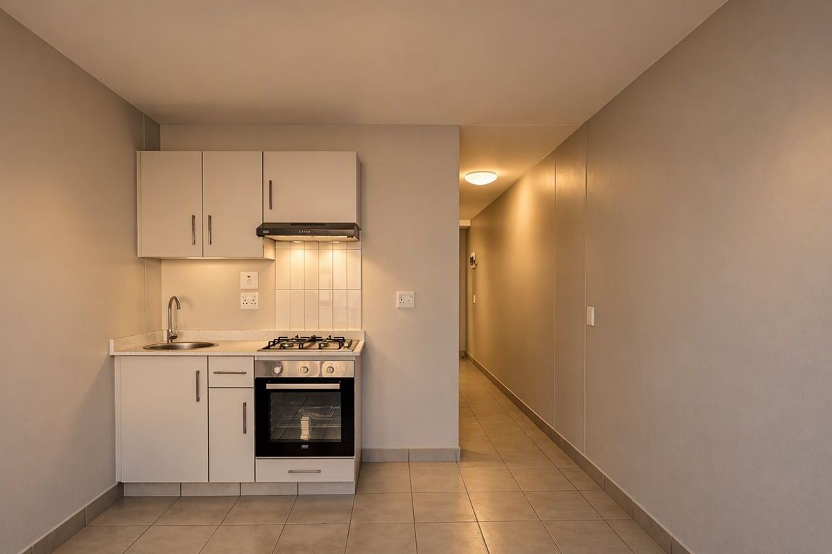 Compact kitchen featuring white cabinetry, built-in oven, gas hob, extractor fan, and tiled splashback, leading into a tiled hallway.