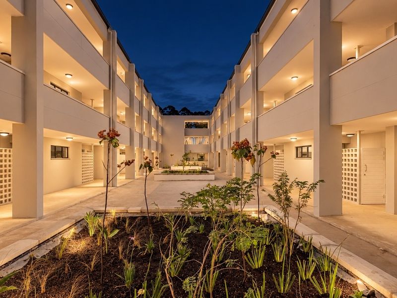 A well-maintained apartment complex courtyard at dusk, featuring illuminated walkways, landscaped garden beds, and modern architectural design.