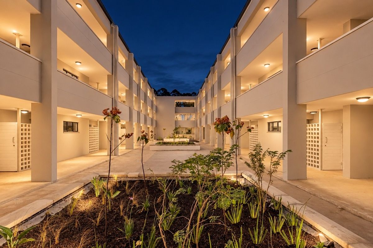 A well-maintained apartment complex courtyard at dusk, featuring illuminated walkways, landscaped garden beds, and modern architectural design.