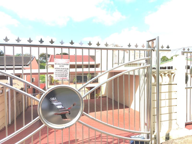 Front view of the property featuring a secure stainless steel gate, paved driveway, and perimeter wall with security fencing.