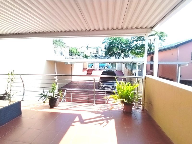 A covered balcony featuring tiled flooring and a stainless steel balustrade, overlooking the driveway and front entrance area.
