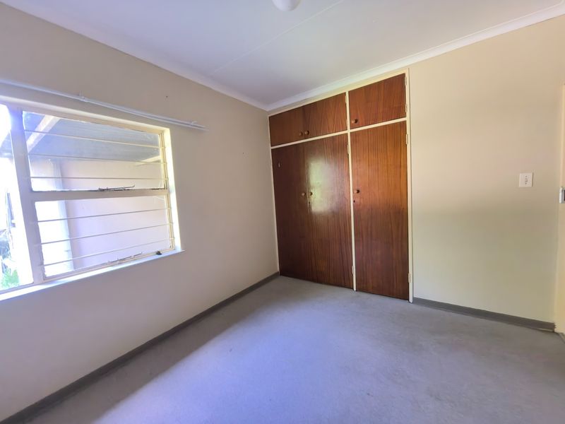 A vacant bedroom featuring built-in wooden cupboards, carpeted flooring, and a window fitted with security burglar bars.