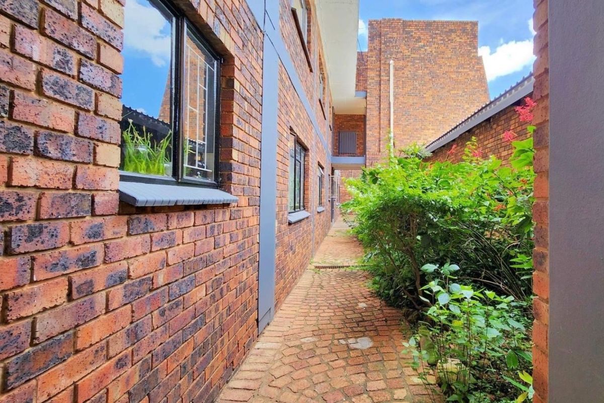 A side exterior walkway featuring face brick walls, aluminum window frames, and a paved path bordered by established garden greenery.