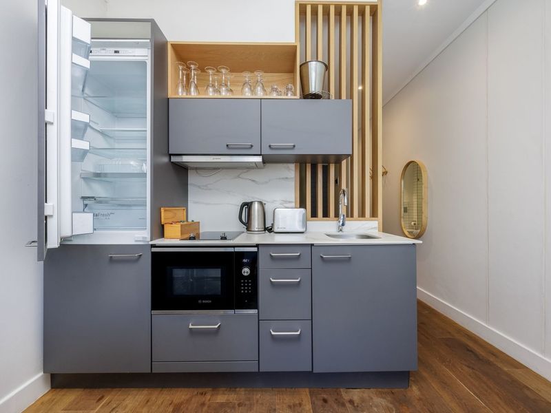 Modern compact kitchen featuring grey cabinetry, integrated appliances, stone countertops, and a decorative timber slat divider, set against hardwood flooring.