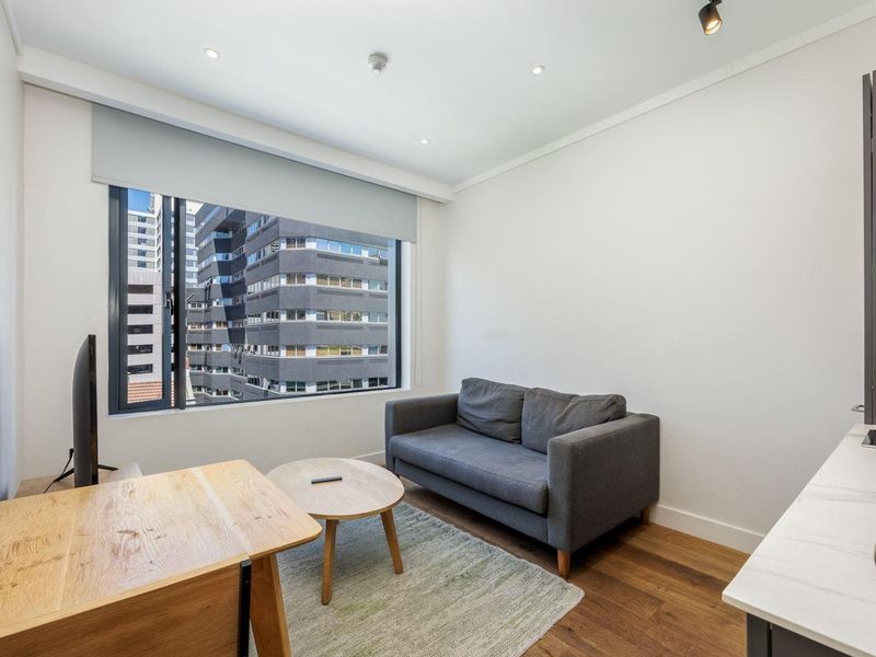 A modern living area featuring timber flooring, recessed downlights, and a large window with urban views, integrated with a compact kitchenette.