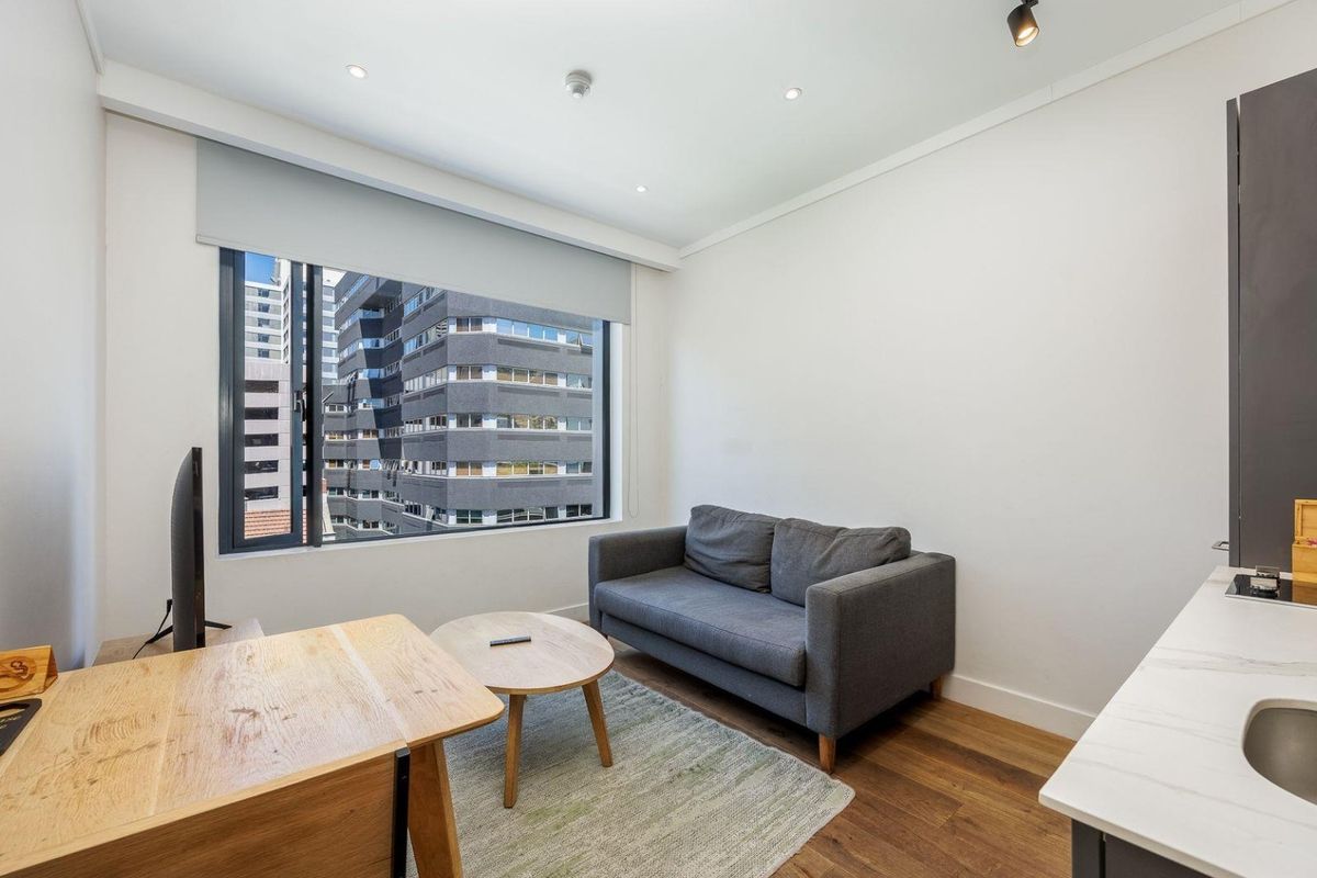 A modern living area featuring timber flooring, recessed downlights, and a large window with urban views, integrated with a compact kitchenette.