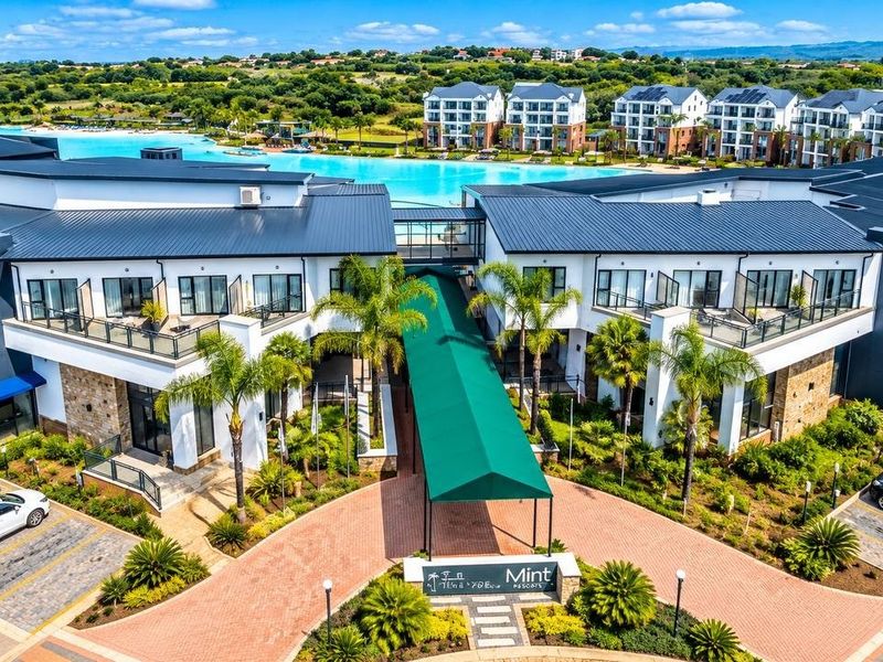 Aerial view of a modern apartment complex featuring a paved entrance, covered walkway, landscaped gardens, and a large swimming lagoon.