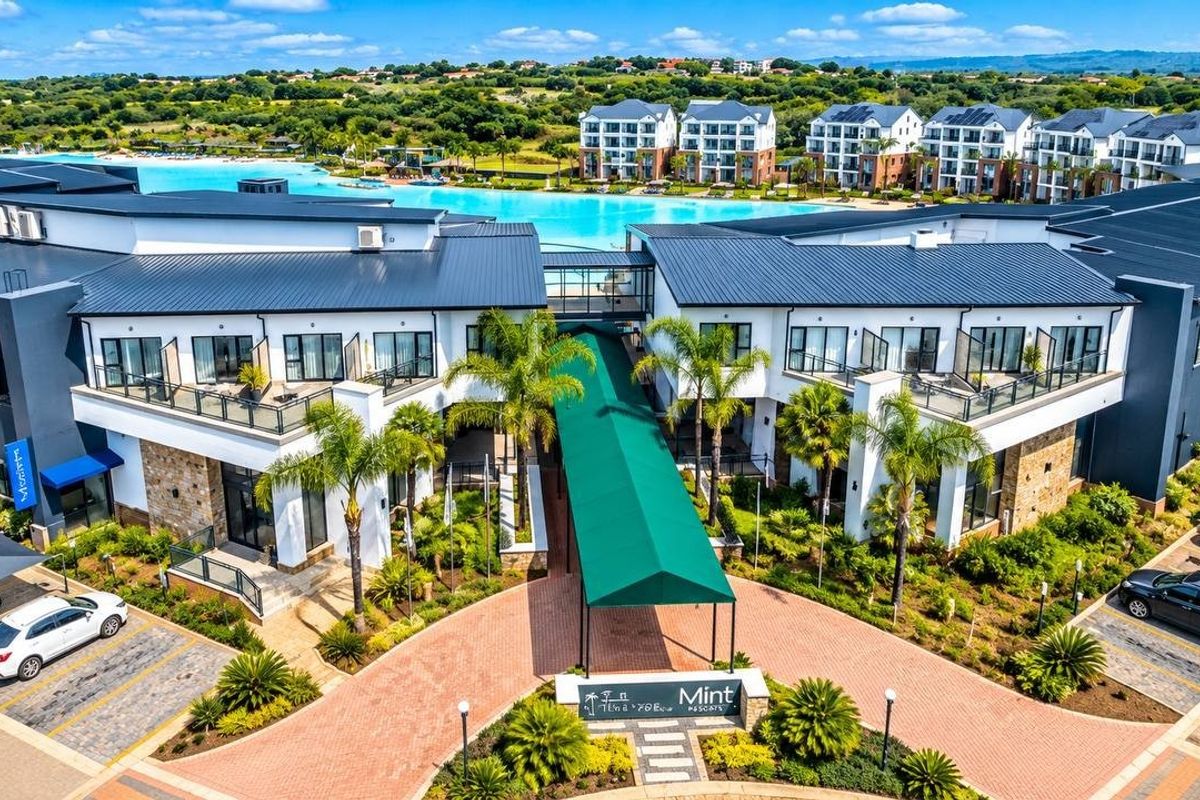 Aerial view of a modern apartment complex featuring a paved entrance, covered walkway, landscaped gardens, and a large swimming lagoon.