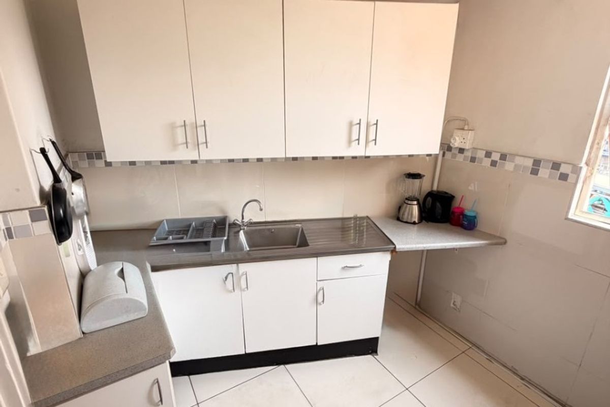 A compact kitchen featuring white built-in cabinetry, a stainless steel sink, tiled backsplash, and neutral floor tiles, offering a functional space for daily use.