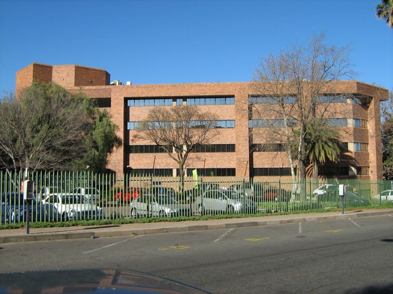 A multi-story commercial office building featuring a classic brick facade, tinted windows, and secure perimeter fencing with an adjacent parking area.
