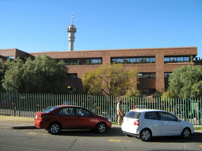 A multi-story commercial office building featuring a classic brick facade, secure perimeter fencing, and tinted windows, situated along a street front.