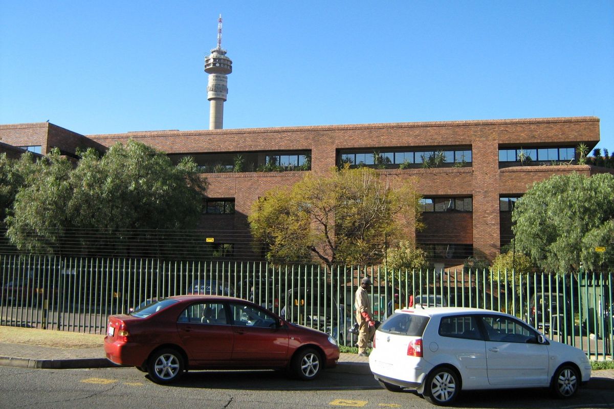 A multi-story commercial office building featuring a classic brick facade, secure perimeter fencing, and tinted windows, situated along a street front.