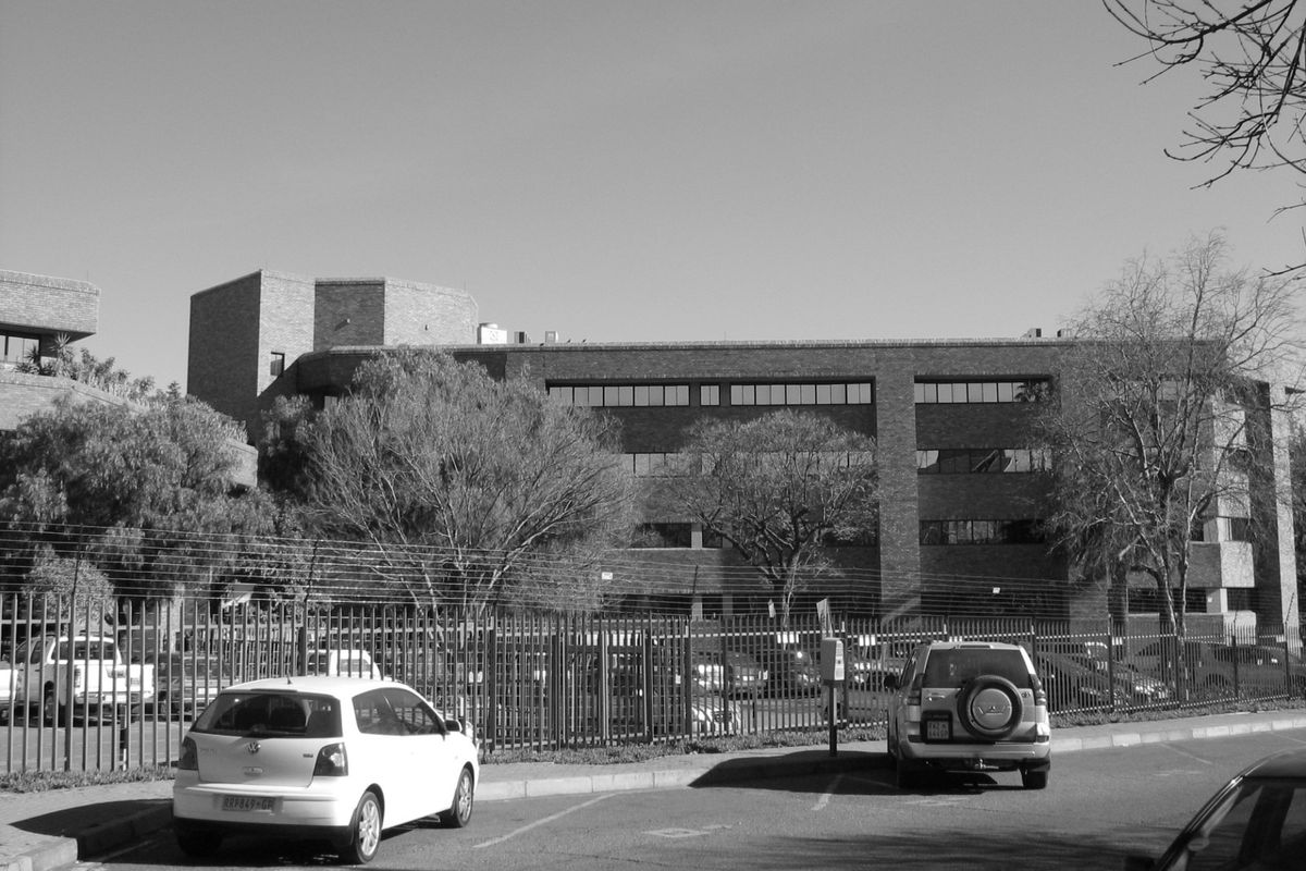 A multi-story commercial office building featuring a brick facade, secured by perimeter fencing with an adjacent paved parking area.