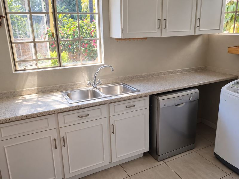 A functional kitchen area featuring white cabinetry, a double basin sink, laminate countertops, and tiled flooring with natural light from the window.