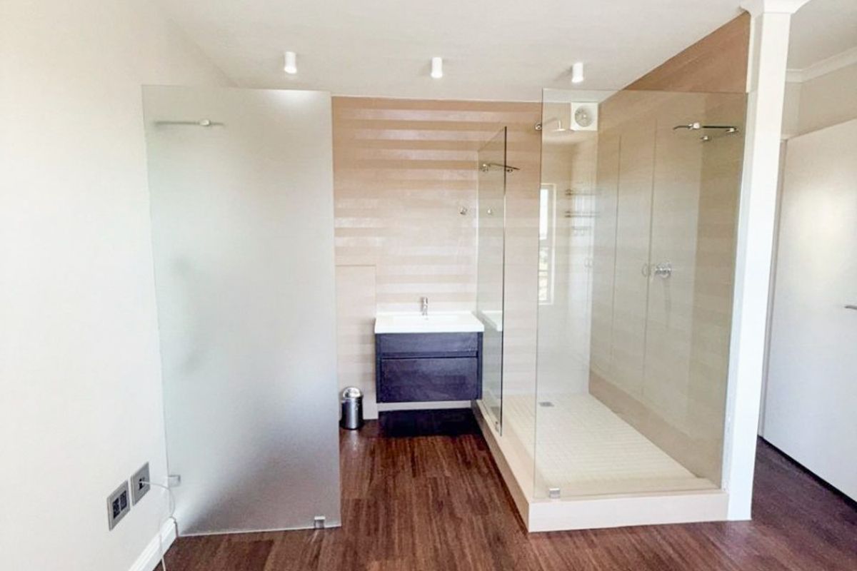 Modern open-plan bathroom featuring a glass-enclosed shower, vanity basin, and dark laminate flooring in a clean, well-maintained space.