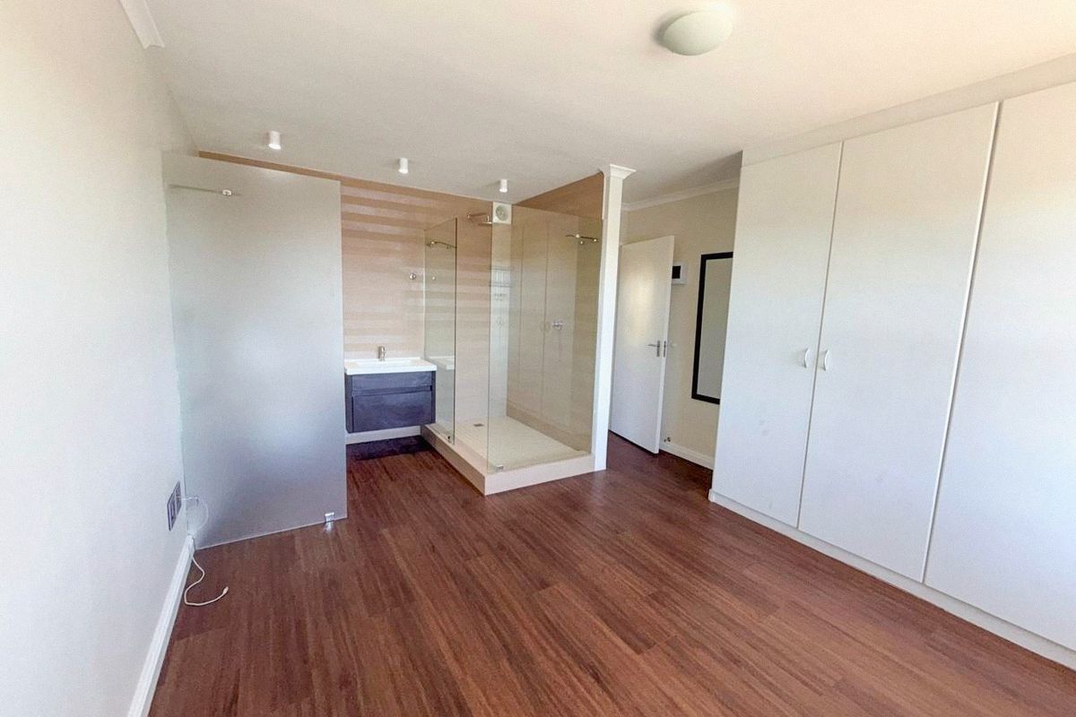 A spacious bedroom featuring integrated built-in cupboards, laminate flooring, and an open-plan en-suite bathroom with a modern glass shower and vanity basin.